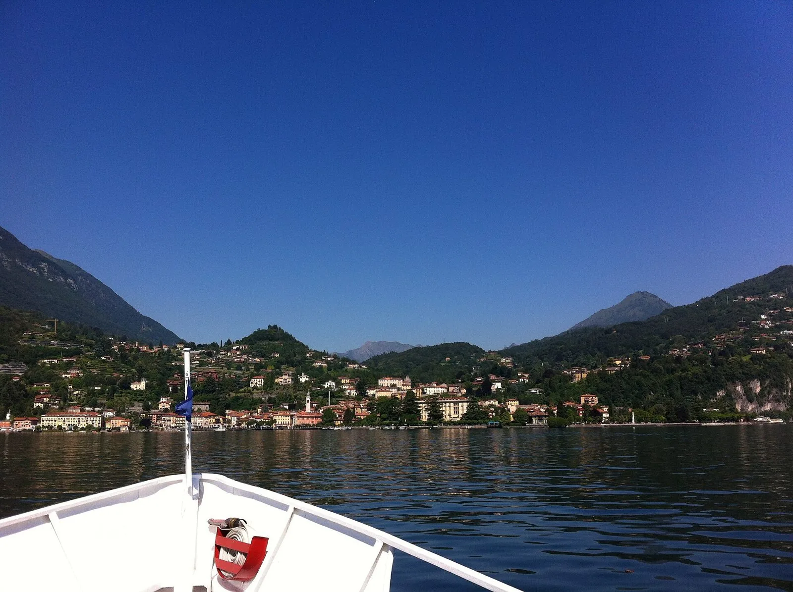 Il Lago di Como visto dalla prua di un battello in una giornata di sole, con un borgo sulla riva e le montagne